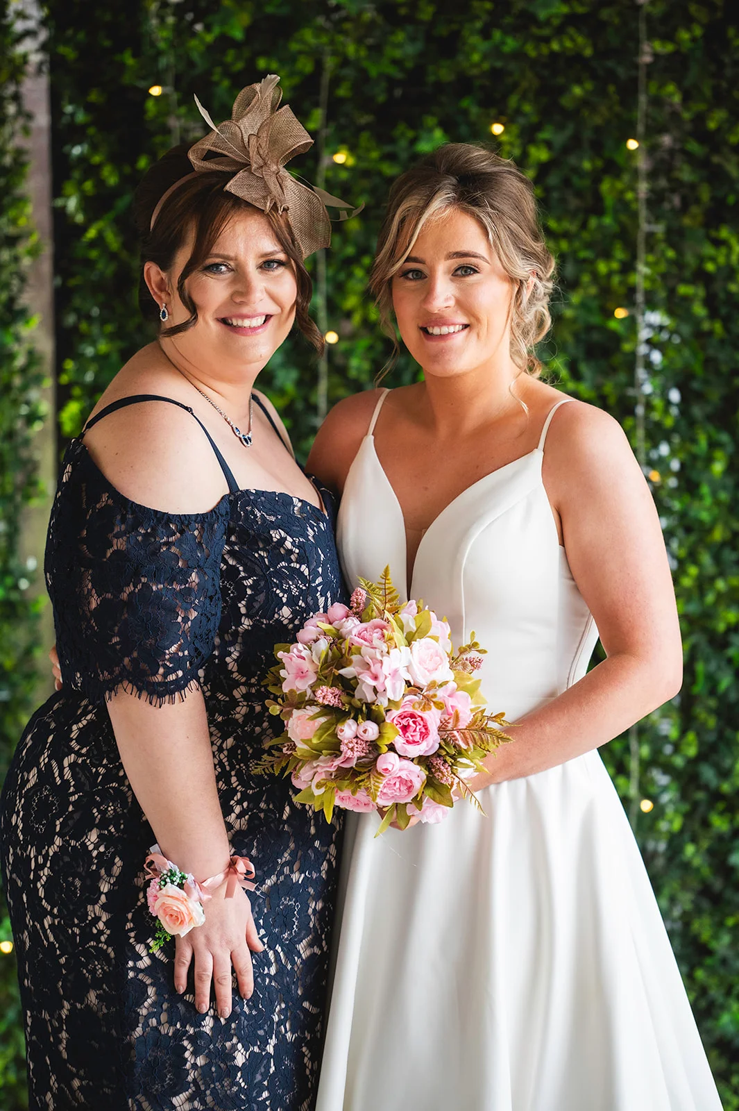 A smiling bride in a white a line gown holding a pink bouquet poses alongside her mother wearing a navy lace dress and a matching fascinator.