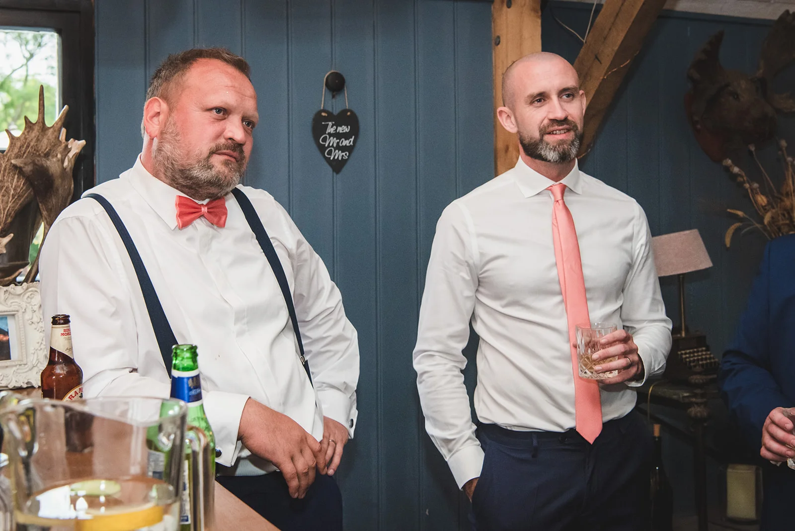 Two well-dressed groomsmen in white shirts and trousers, one wearing braces and a peach bow tie and the other a peach necktie, listen attentively during the wedding speeches.