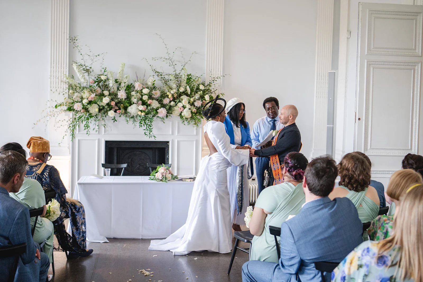 An interracial couple holding hands during a civil ceremony in front of a white fireplace adorned with a large floral installation.