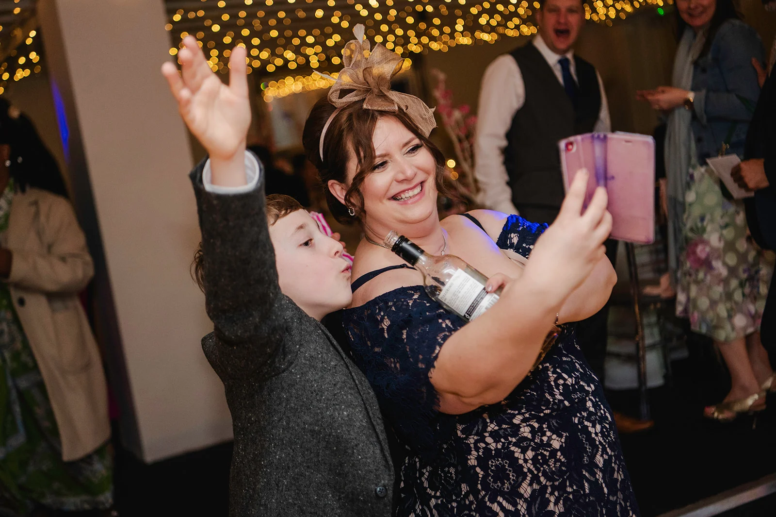 A joyful wedding guest wearing a fascinator and navy lace dress takes a selfie on her phone while holding a bottle, accompanied by a young boy raising his hand excitedly beneath a canopy of fairy lights.