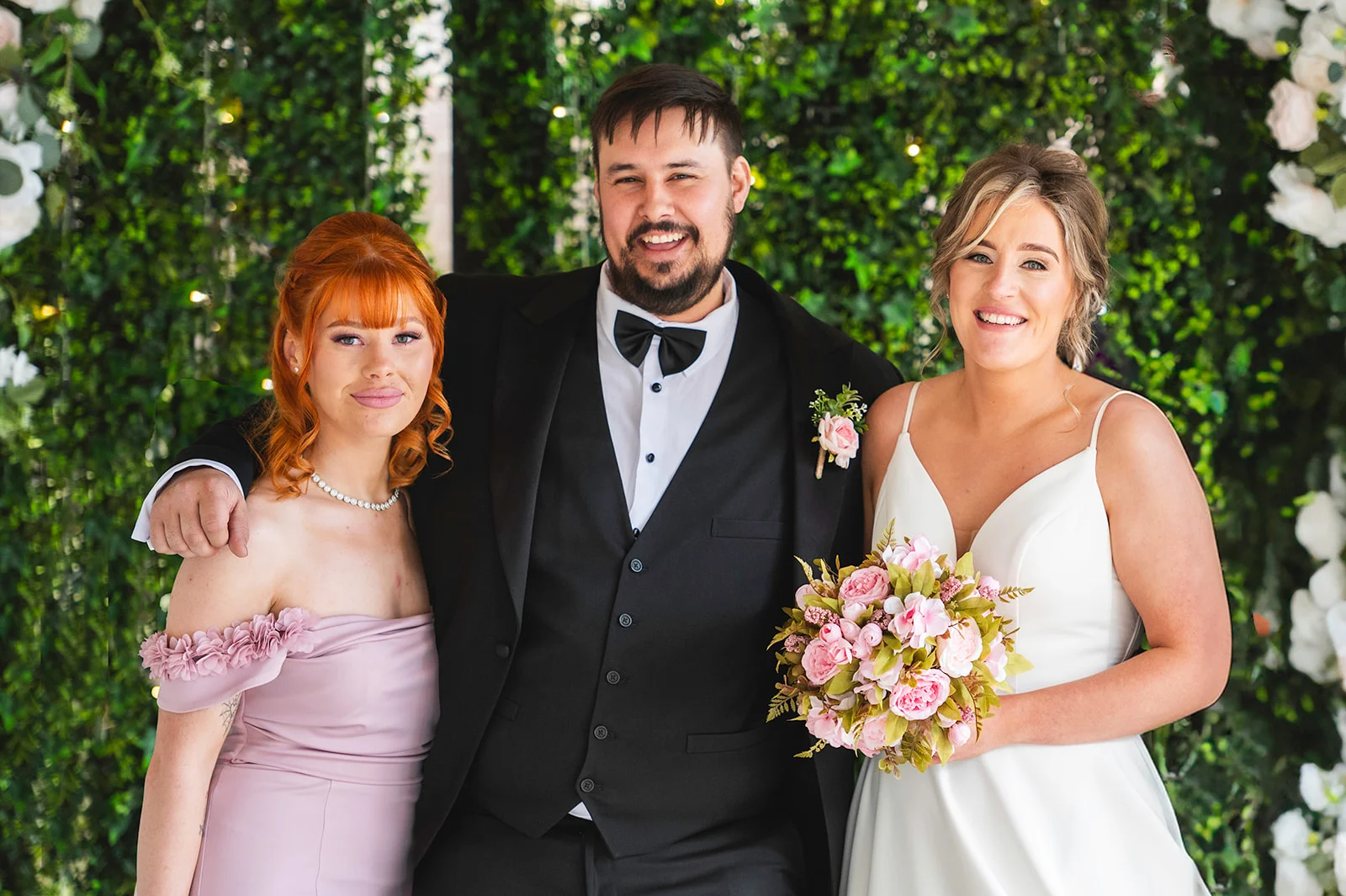A smiling groom in a black tuxedo stands between a bridesmaid in a pink off-the-shoulder dress and a bride holding a pink bouquet in front of a lush greenery wall.