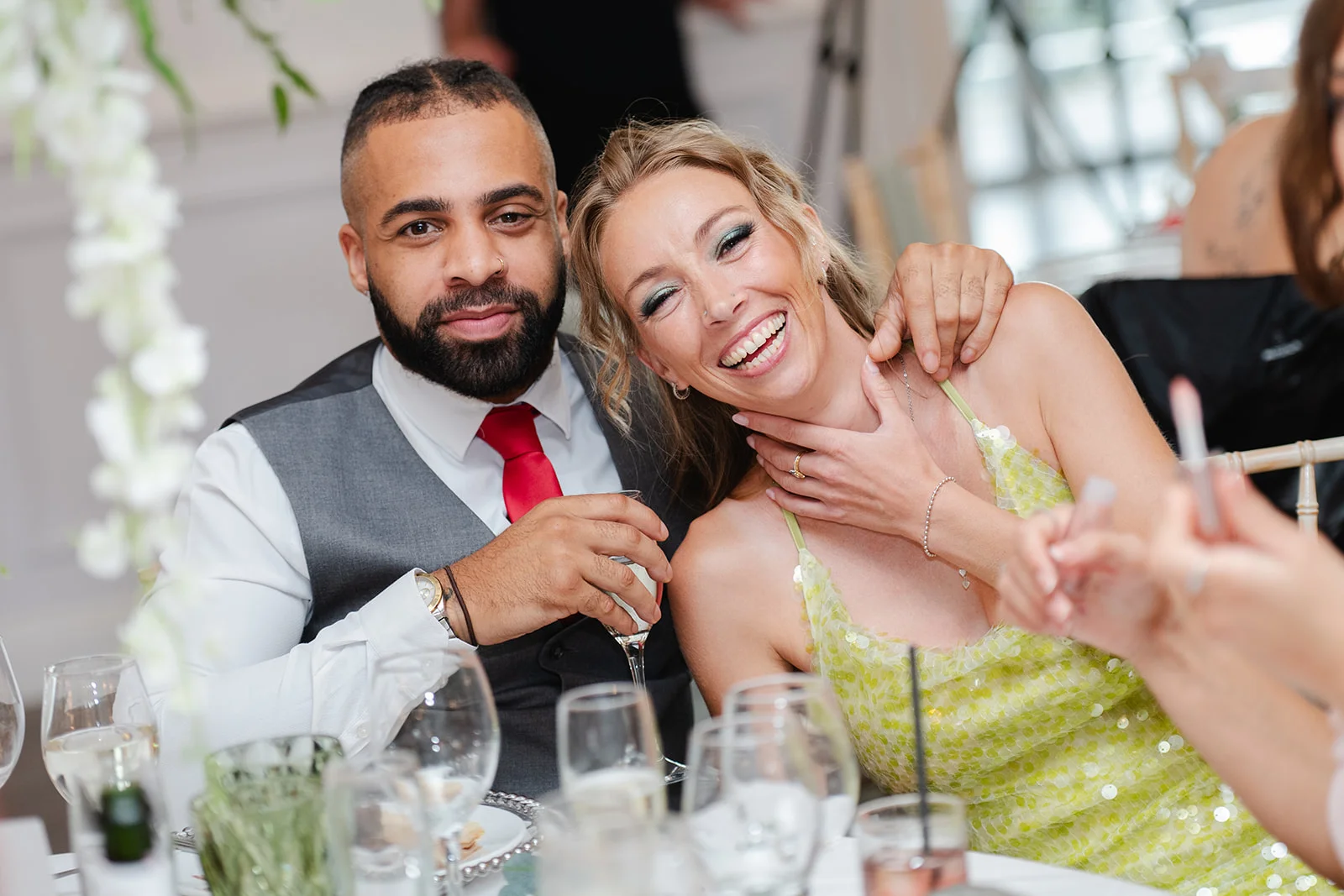 A joyful newlywed couple smiling and laughing together at a wedding reception table while holding champagne glasses.