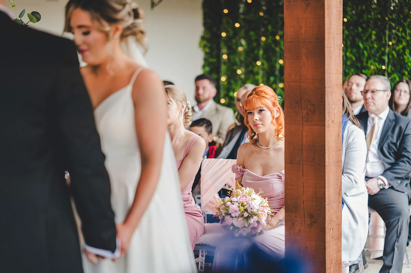 A candid wedding ceremony shot capturing a bridesmaid in a pink dress holding a bouquet while watching the couple at the altar.