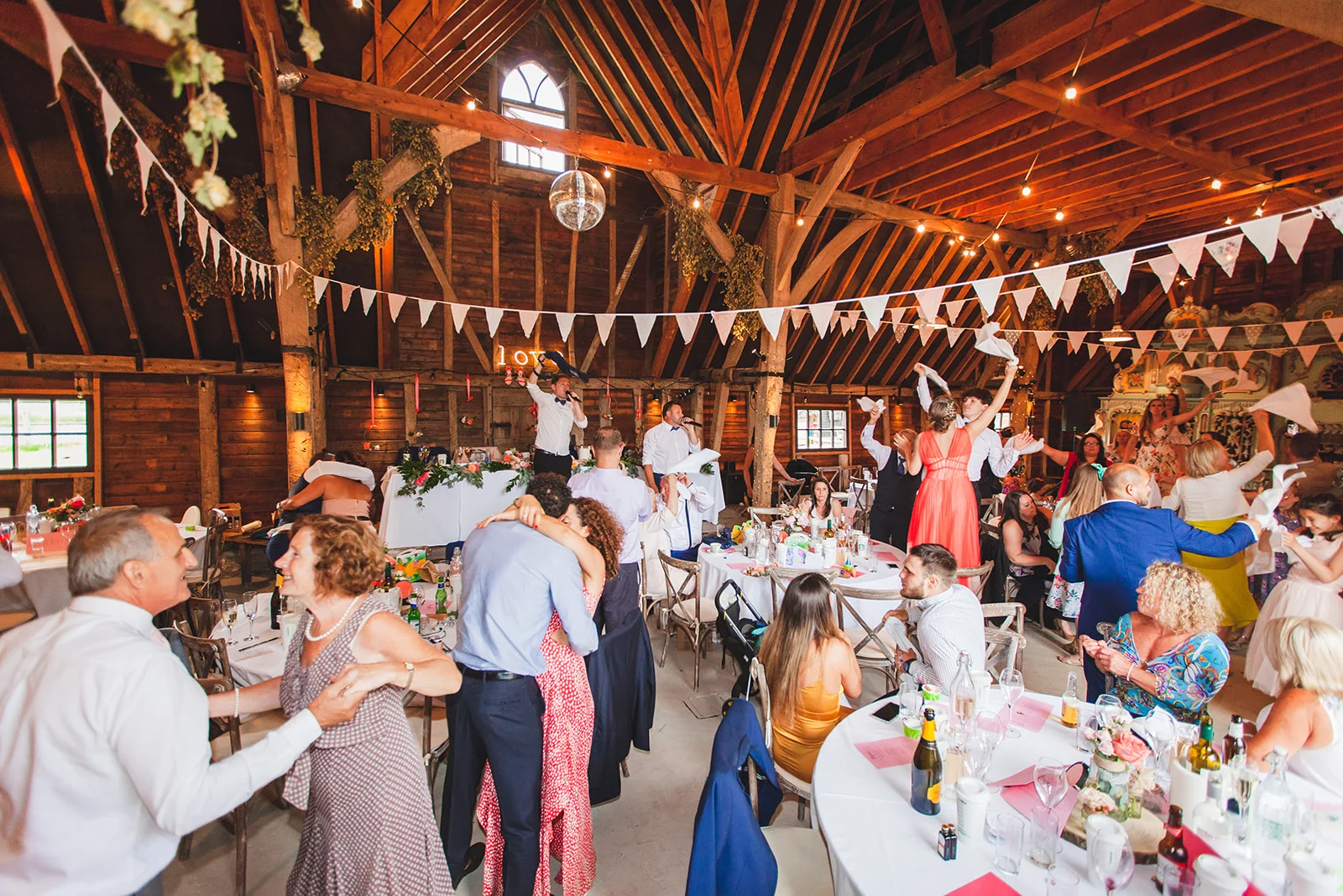 Lively wedding reception inside a rustic oak-framed barn with guests celebrating beneath exposed rafters and hanging bunting.