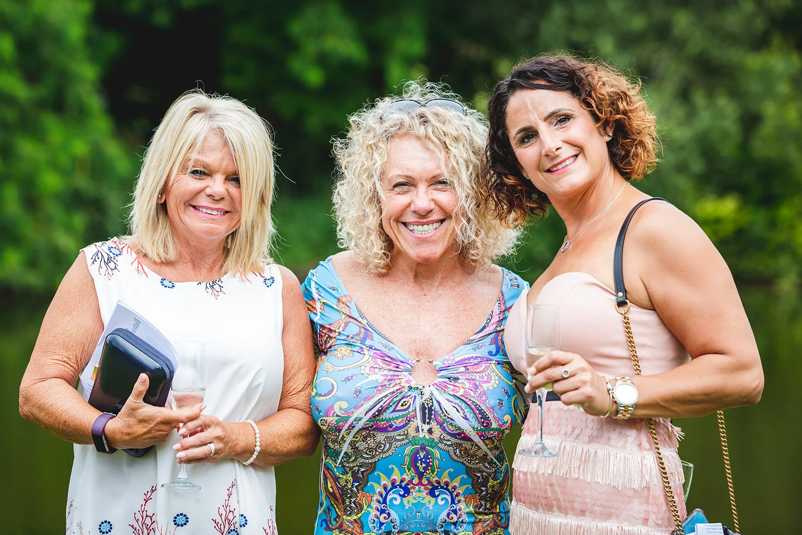 Three female wedding guests smiling together outdoors while holding champagne glasses during a drinks reception.