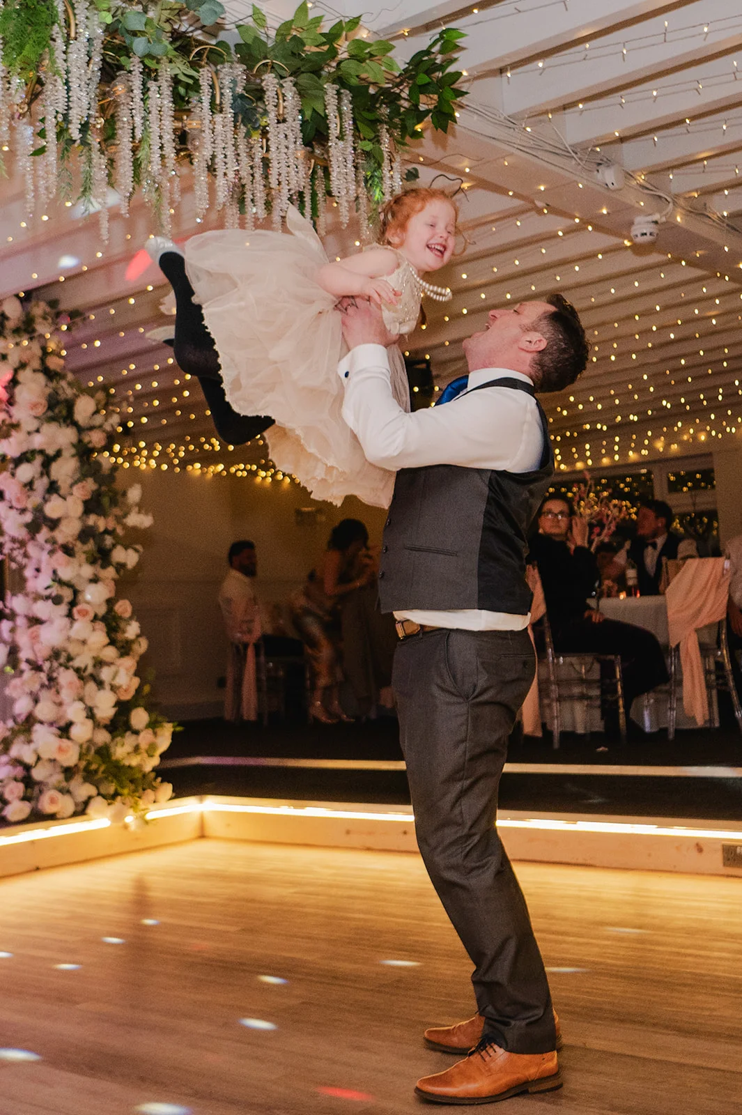 A joyful wedding guest joyfully lifts a smiling young flower girl in a fluffy tulle dress high into the air on a brightly lit wooden dance floor.