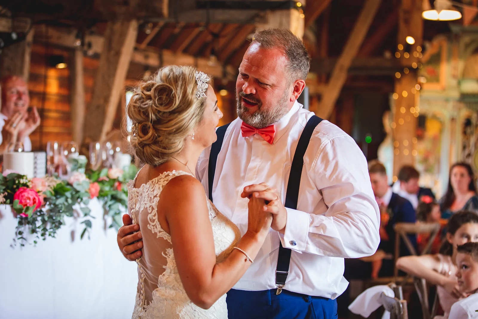 The bride and groom share a joyful first dance together inside a rustic barn venue decorated with exposed wooden beams and floral arrangements.