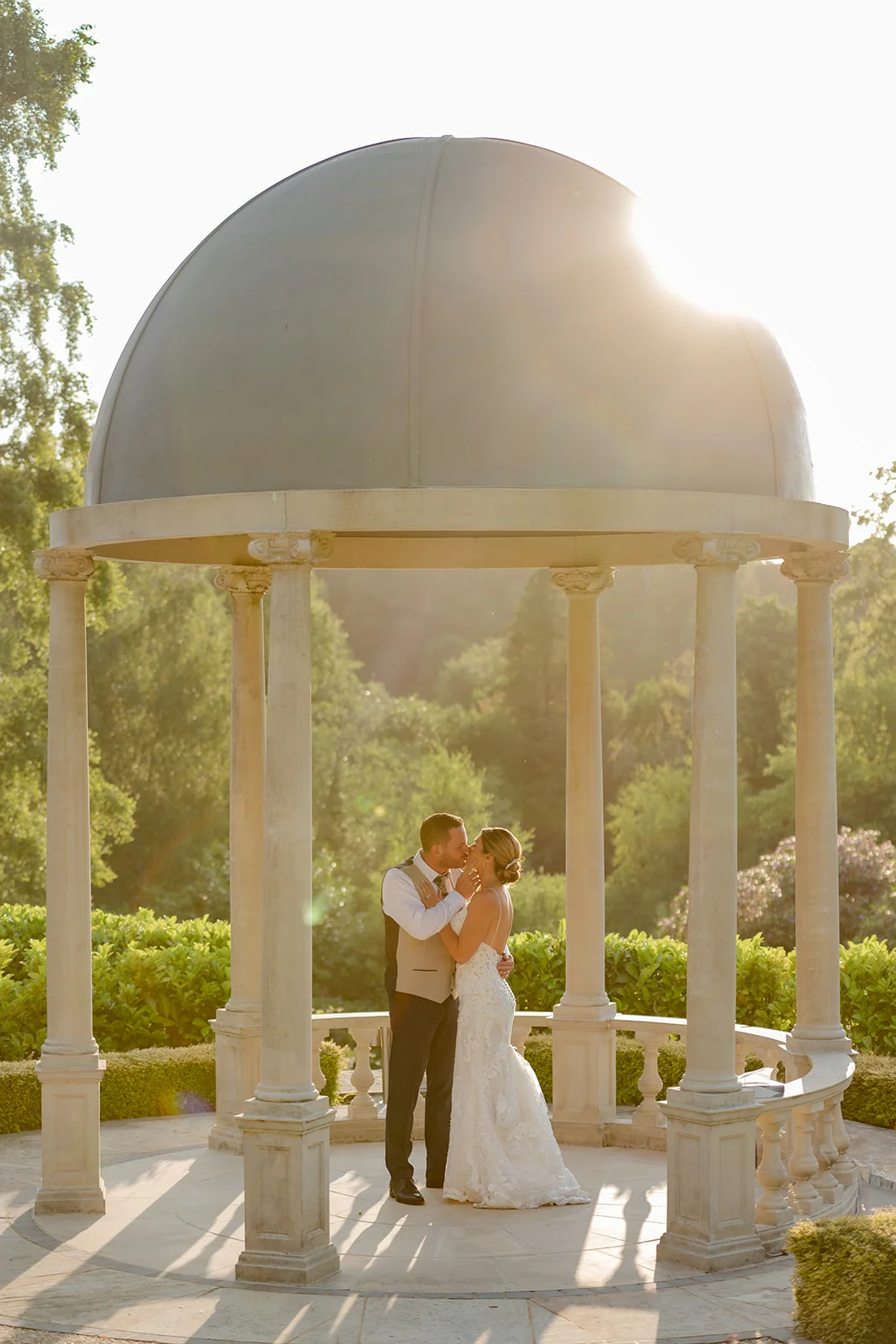 A newlywed couple shares a tender kiss inside an ornate stone gazebo bathed in warm, glowing golden hour sunlight.