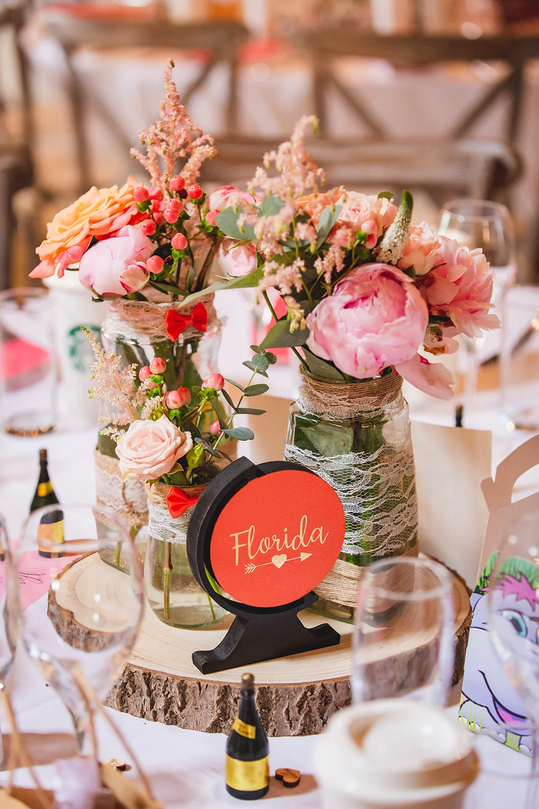 A rustic wedding table centrepiece featuring pink and peach peonies and roses arranged in lace-wrapped mason jars on a wood slice with a travel-themed table sign.