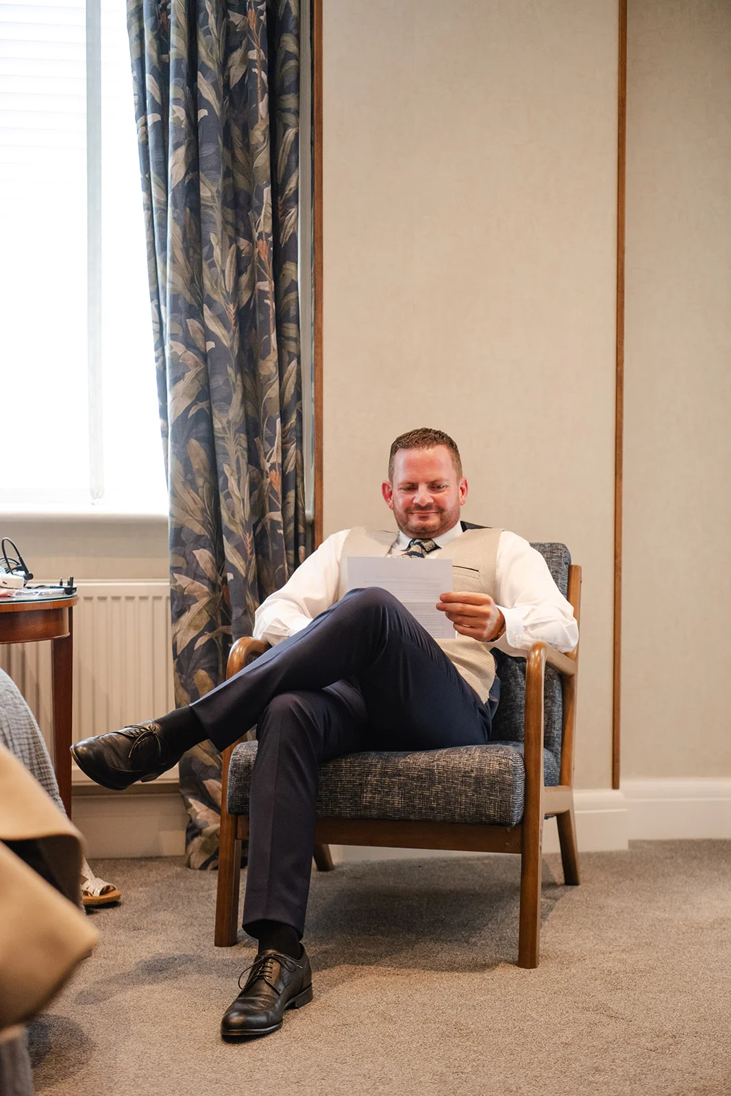 A groom sits comfortably in an armchair near a window, smiling gently as he reads a letter while getting ready for his wedding day.