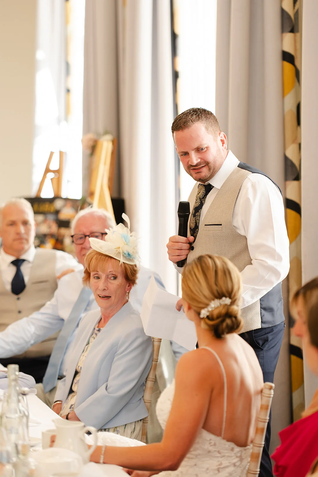 A groom delivers a speech holding a microphone and notes at the top table while seated guests look on during the wedding breakfast.