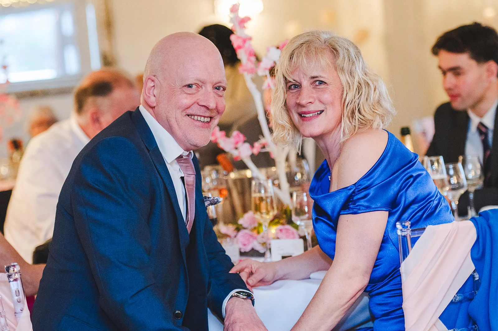 An older couple smiling happily together at a wedding reception table set with floral centrepieces and wine glasses.