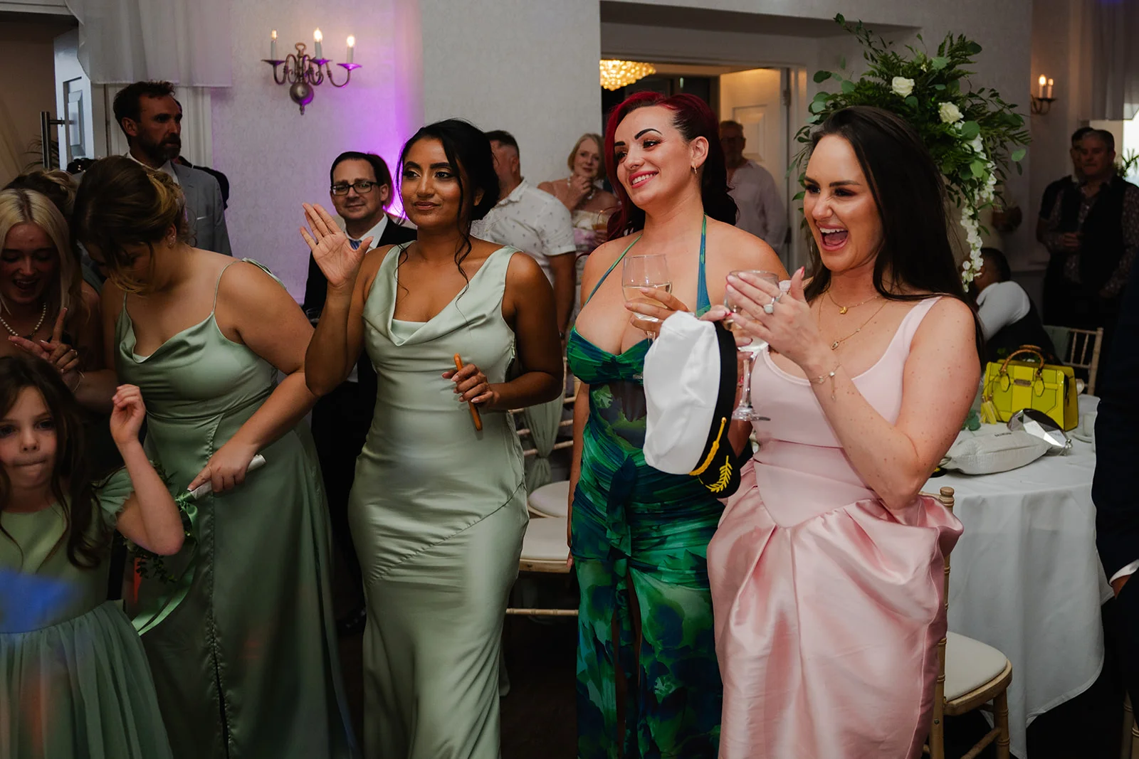 A joyful group of bridesmaids and wedding guests laughing and cheering together during an indoor evening reception.