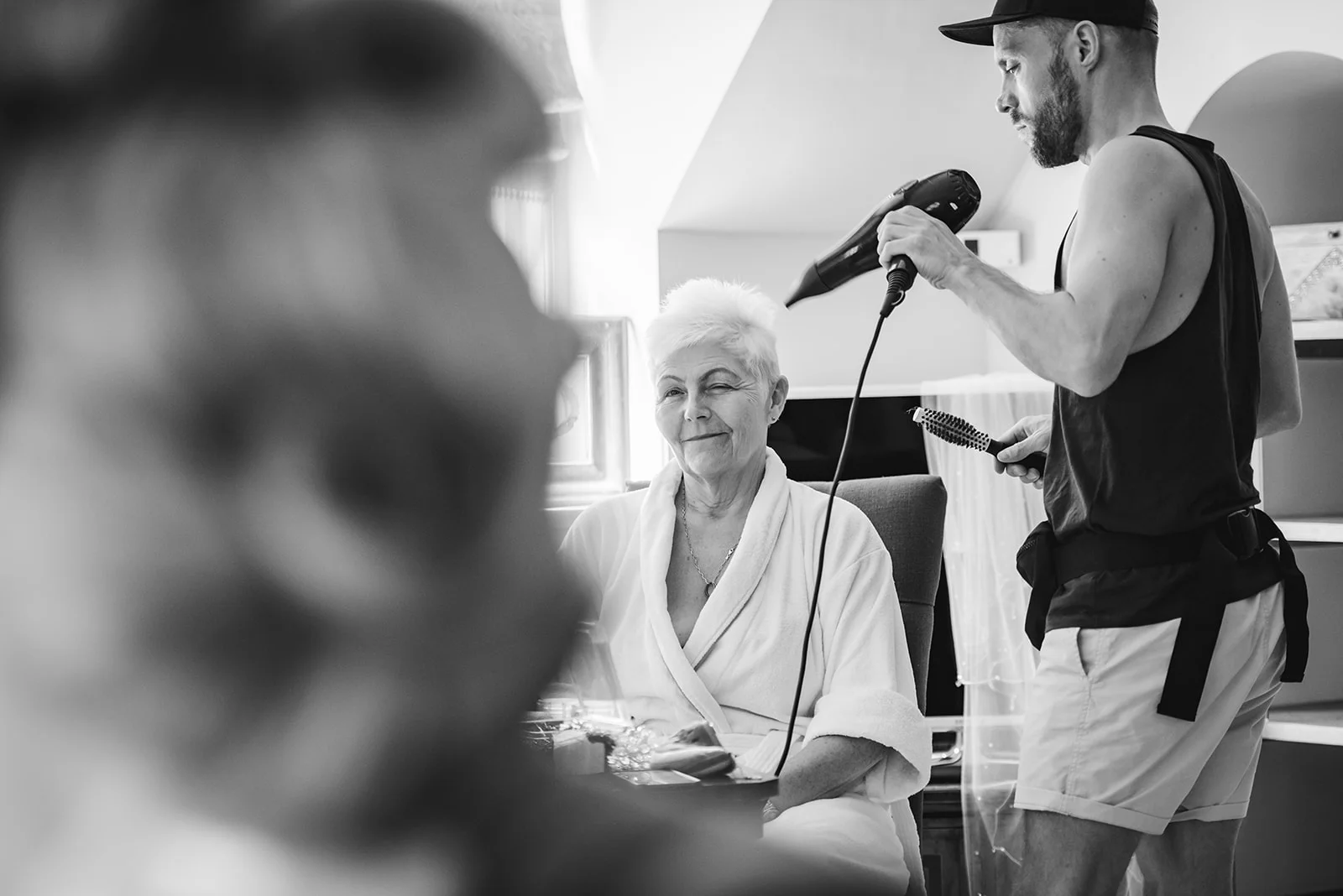 A mature bride wearing a white bathrobe smiles while a stylist blow-dries her hair during wedding morning preparations.
