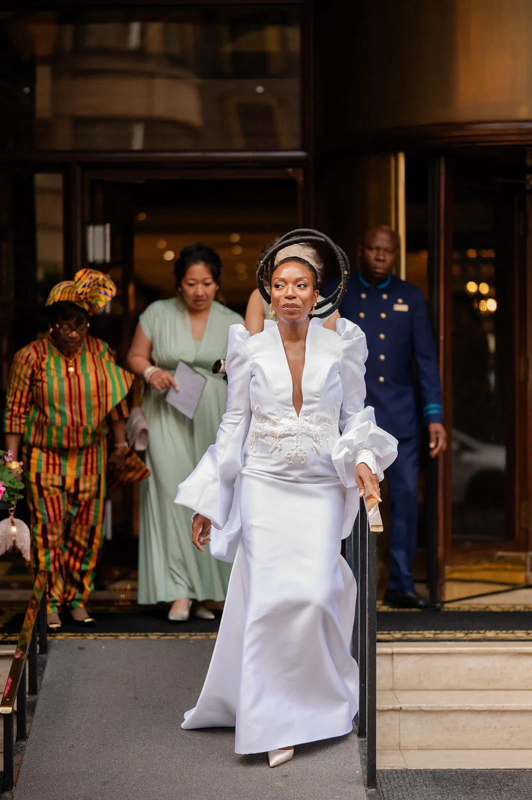 A stunning bride in a white satin gown with dramatic sleeves and a sculptural halo headpiece descends steps outside a hotel entrance.