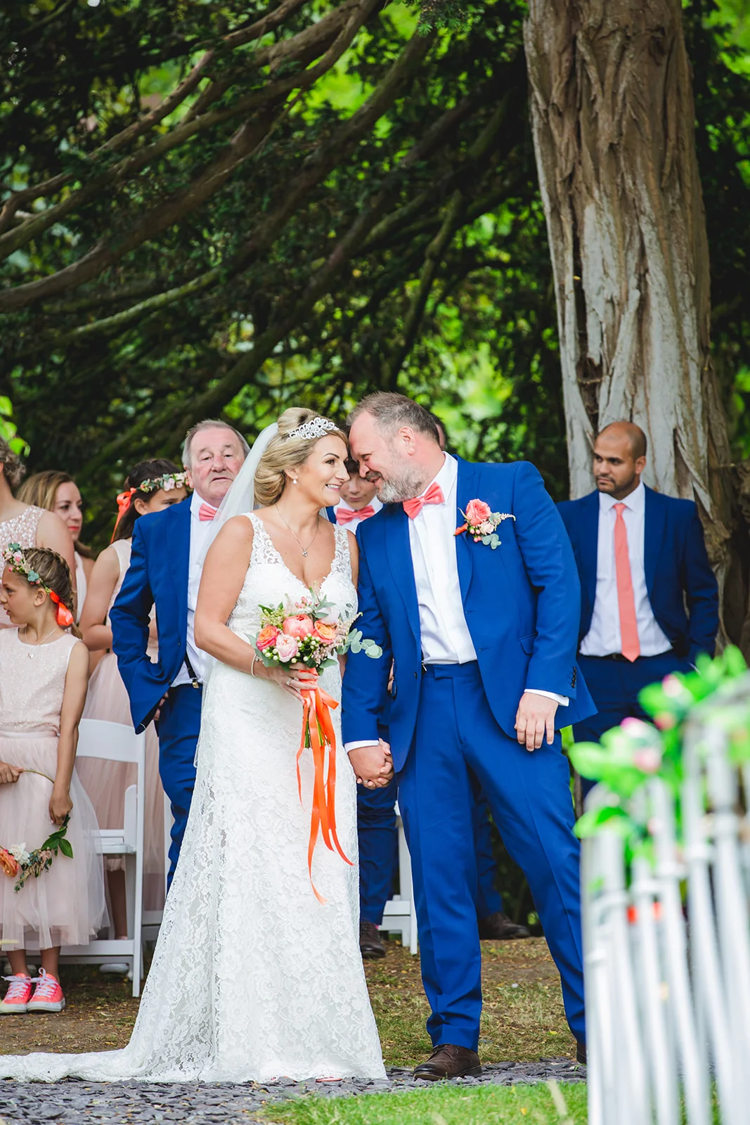 A smiling bride and groom walk hand in hand down the aisle during an outdoor ceremony, surrounded by wedding party members in blue suits and coral accessories beneath mature trees.