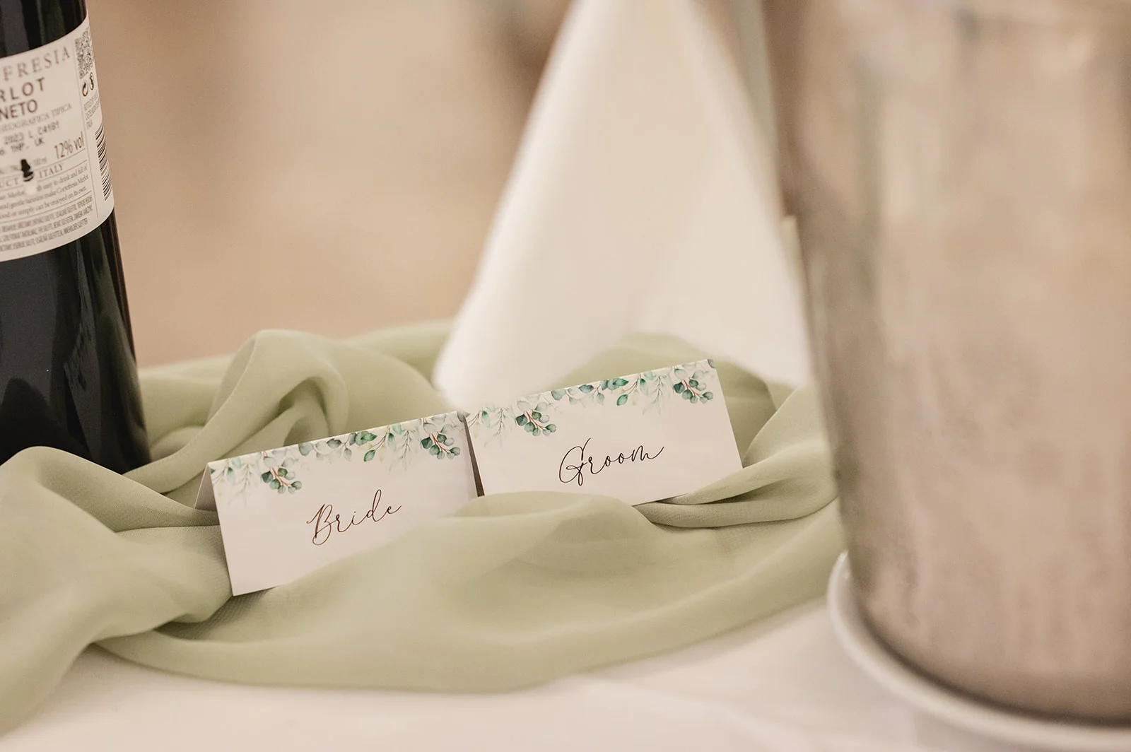 Close-up of elegant place cards labelled 'Bride' and 'Groom' resting on a sage green fabric runner next to a wine bottle.