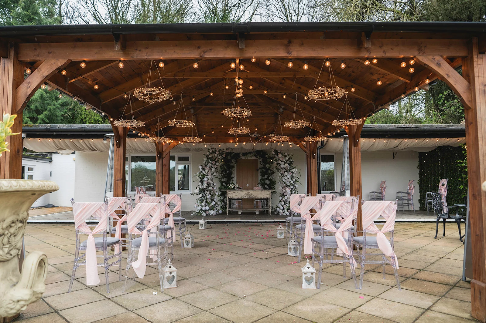 An outdoor wedding ceremony setup features transparent chairs adorned with pink sashes, arranged under a wooden pavilion strung with twinkling fairy lights and hanging floral details.