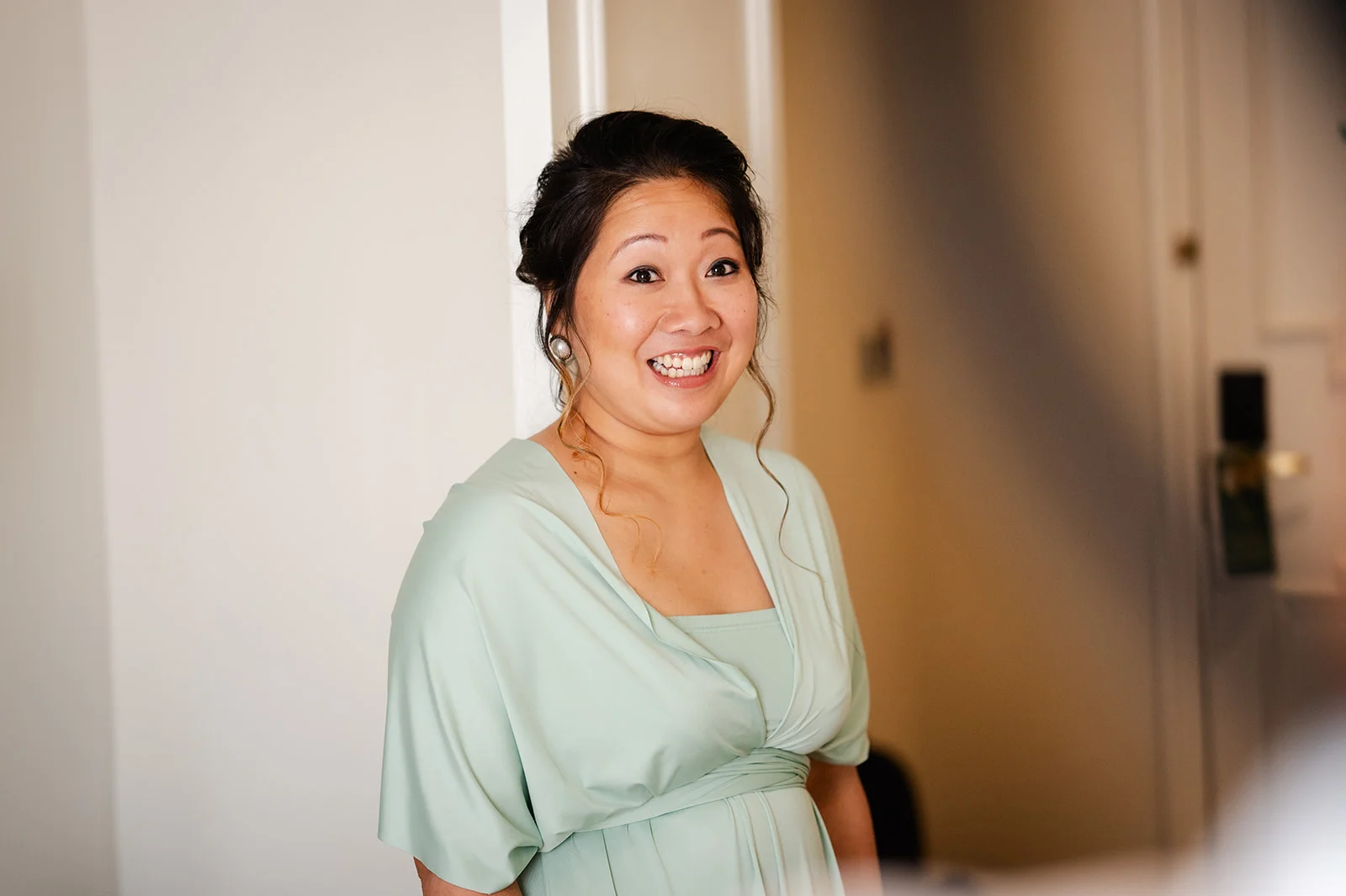 An East Asian bride smiles joyfully while getting ready, wearing a light sage green dress and pearl earrings with her hair in a half-up style.