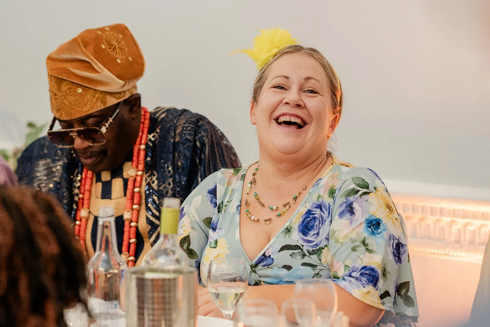 A guest wearing a floral dress and a yellow fascinator laughs heartily during the wedding reception, seated next to a man in traditional attire and beaded necklaces.