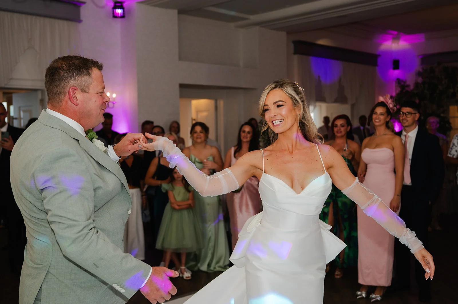 A smiling bride in a satin gown with sheer opera gloves shares a joyful first dance with her groom during the evening reception.