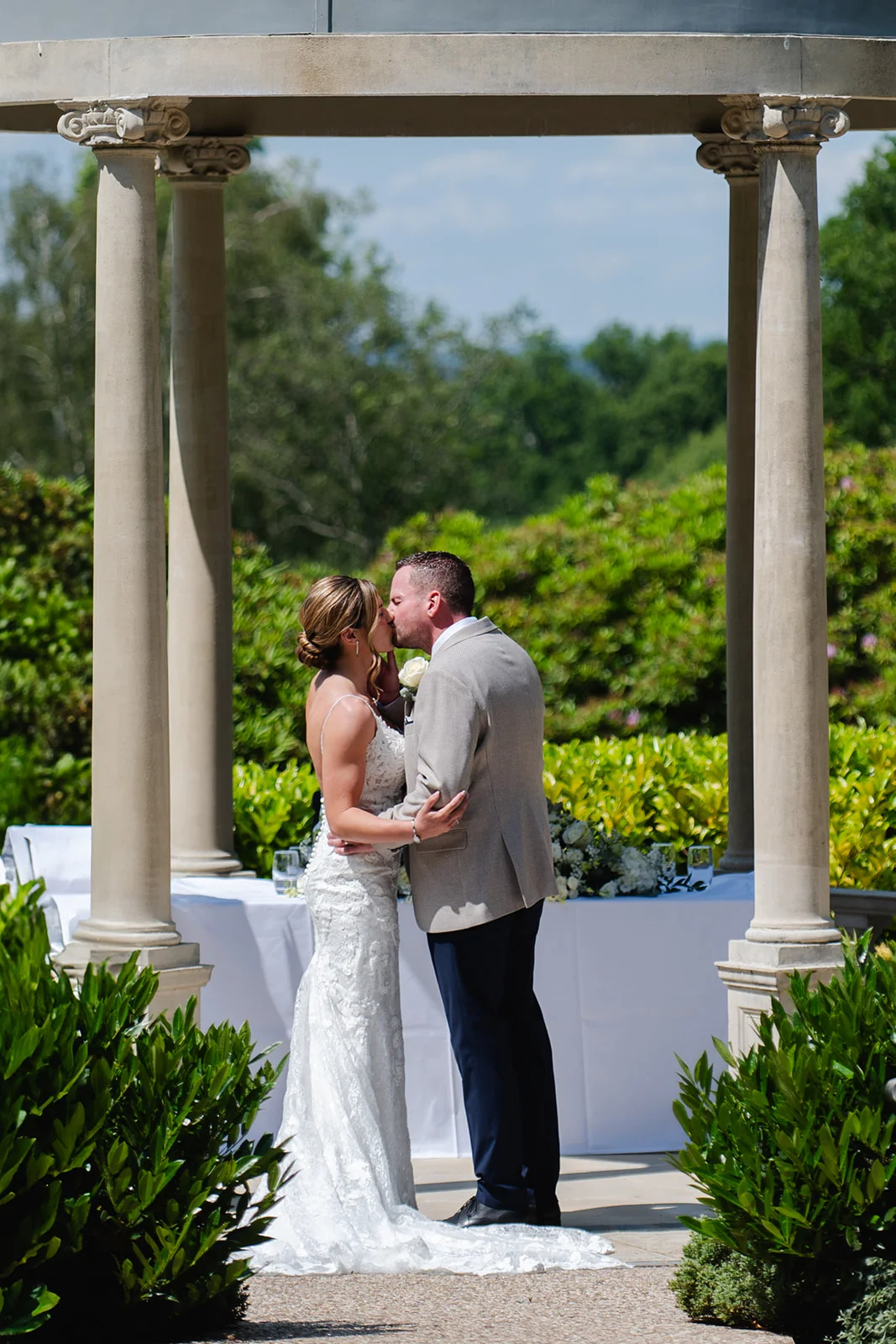A bride and groom share a kiss underneath a stone gazebo during their sunlit outdoor summer wedding ceremony.