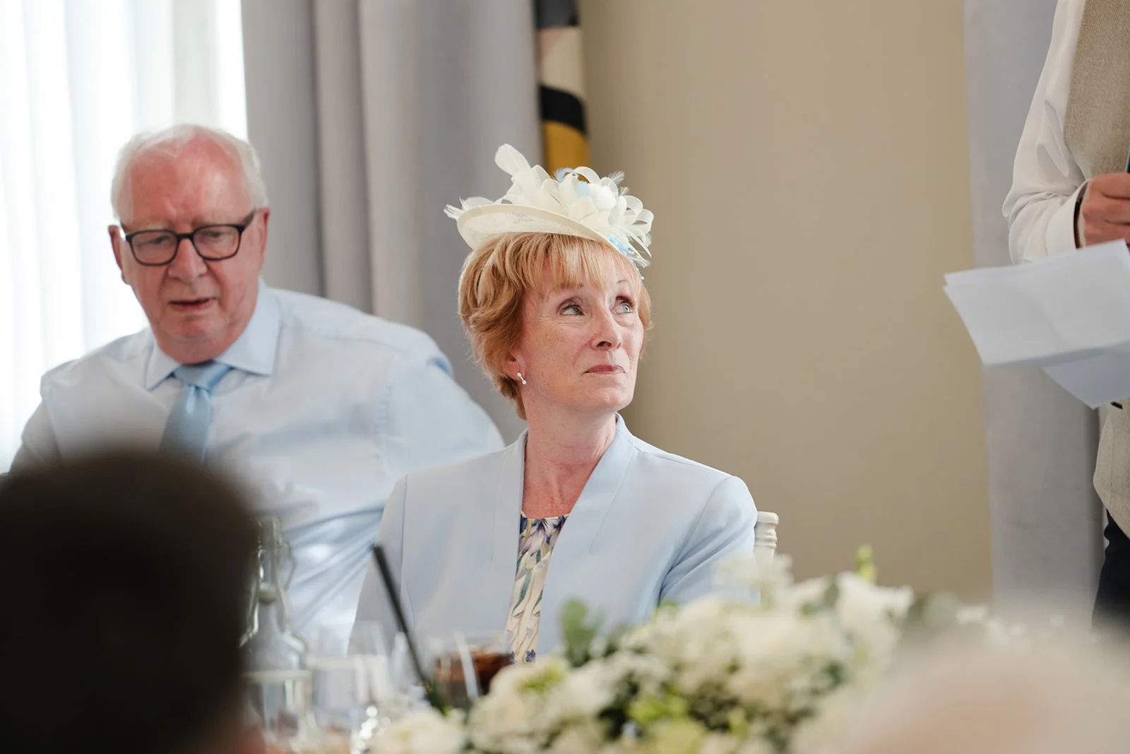 An elegant mother of the bride wearing a pale blue jacket and a decorative fascinator listens emotionally to wedding speeches at the top table.