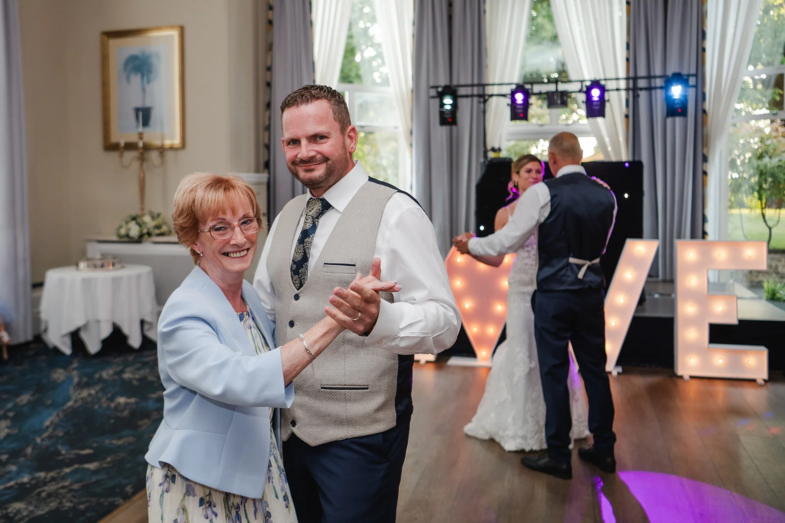 An older woman smiling warmly while dancing with a man in a light grey waistcoat on a wooden dance floor, with another couple dancing in front of illuminated love letters in the background.