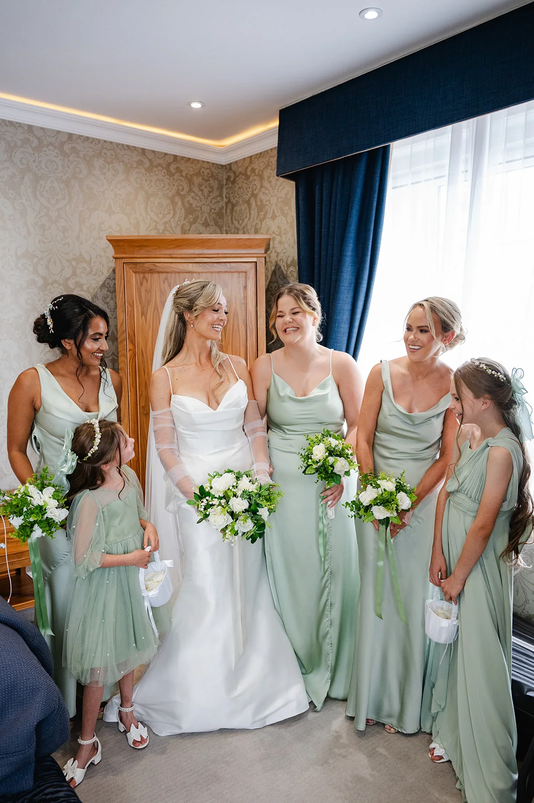 A smiling bride in a satin gown and sheer sleeves stands with her bridesmaids and flower girls dressed in sage green satin gowns, all holding white floral bouquets.