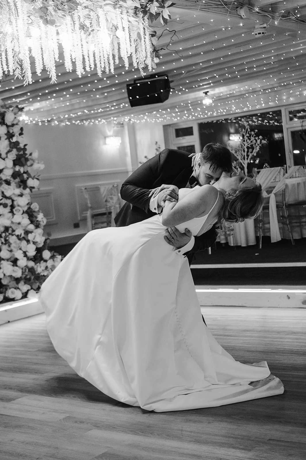 A newlywed couple shares a dramatic dip kiss on the wooden dance floor underneath a sparkling fairy light canopy and floral ceiling installation.
