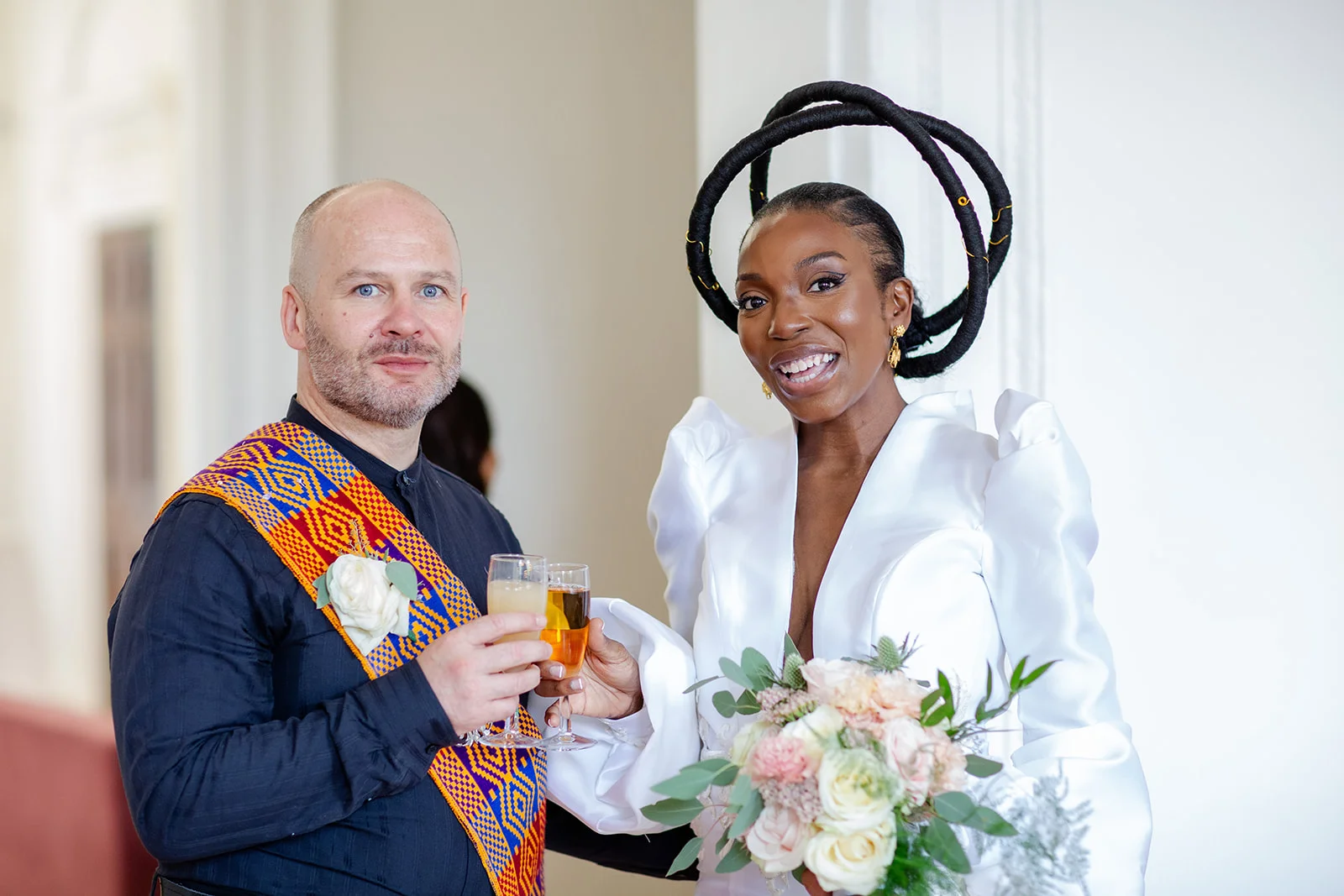 An interracial newlywed couple smiling and clinking glasses of champagne during their wedding reception, featuring a bride in an avant-garde white dress with a sculptural halo headpiece and holding a pastel bouquet.