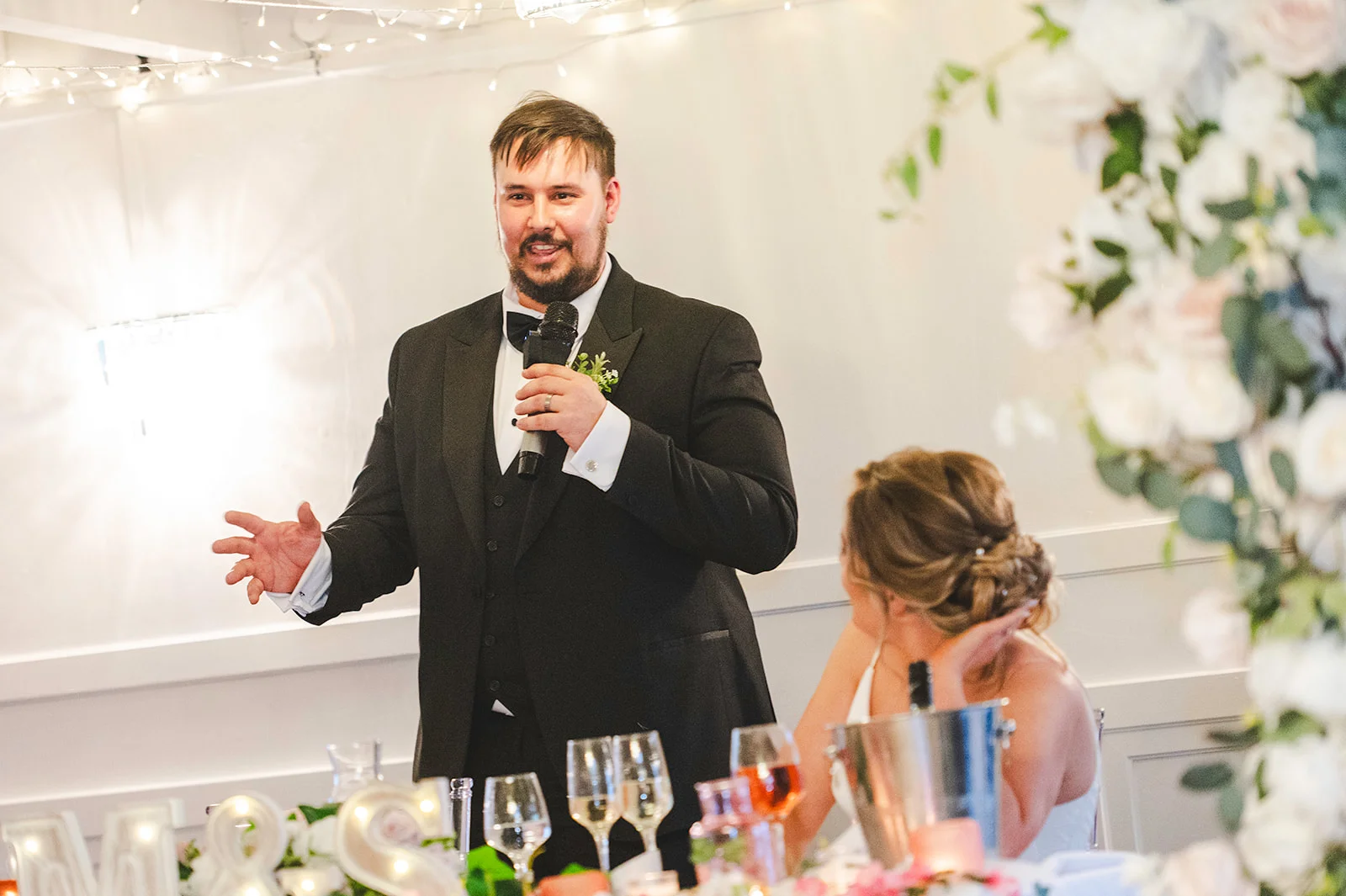 A groom in a classic black tuxedo delivers a speech holding a microphone during the wedding reception while his bride sits nearby at the top table.