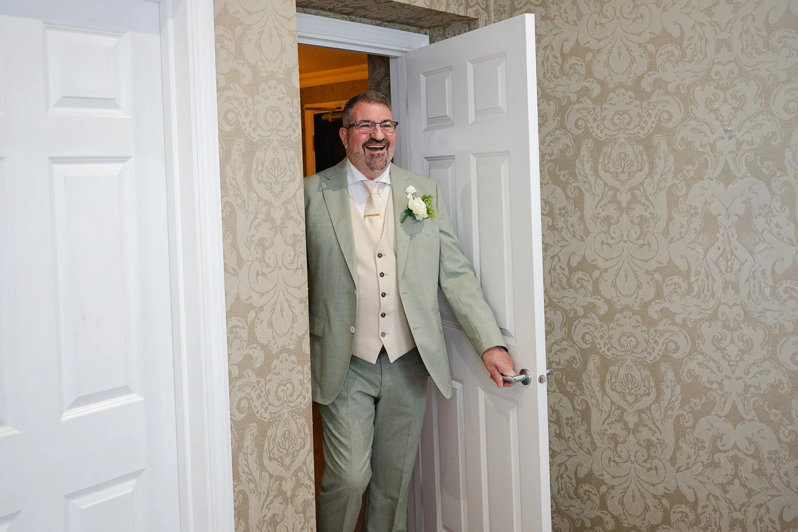 A smiling father of the bride wearing a sage green suit and a matching waistcoat proudly opens a white door during wedding morning preparations.