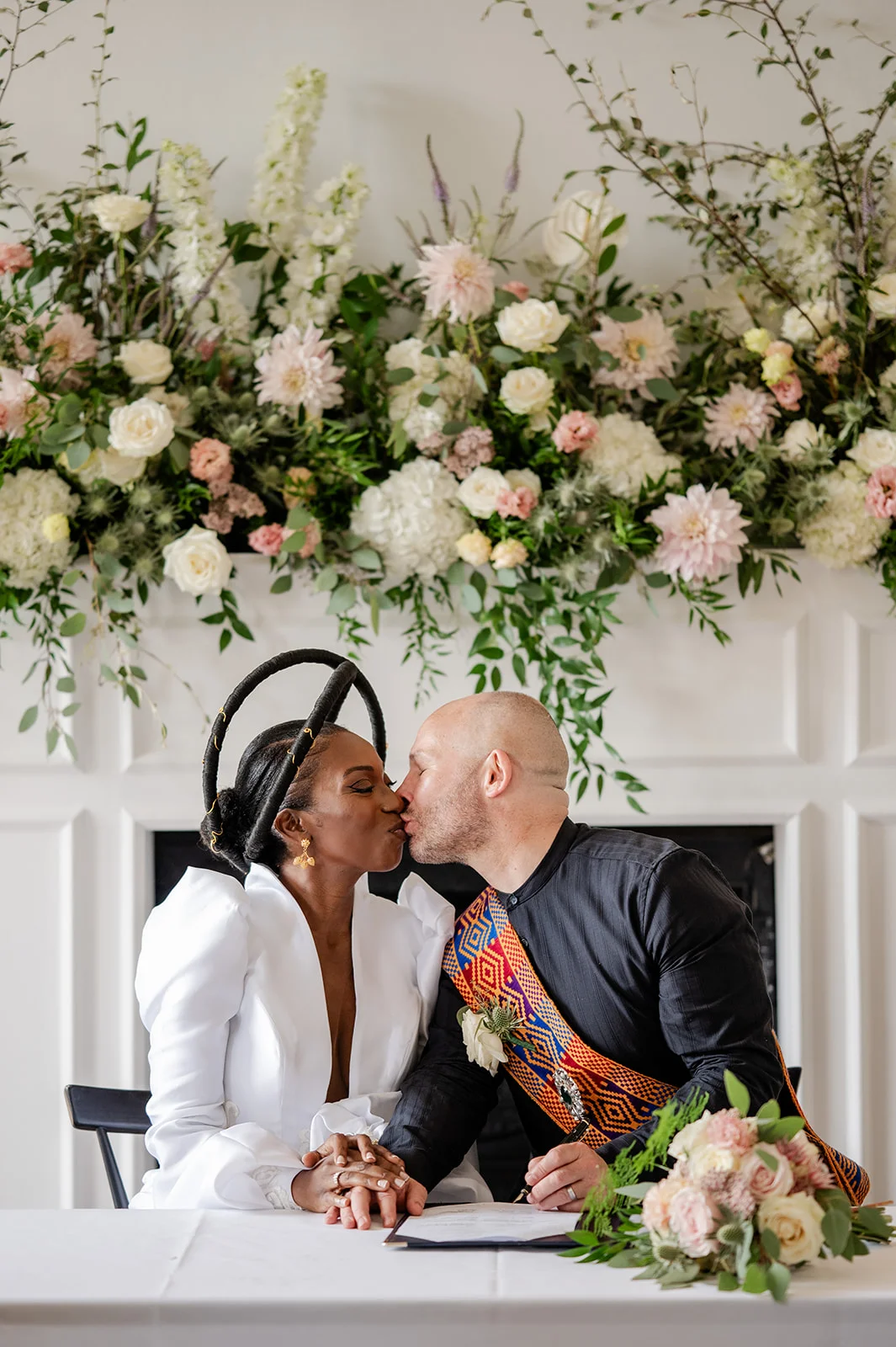 A newly married interracial couple share a tender kiss while holding hands over a wedding certificate in front of a grand floral mantelpiece display.