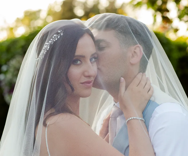 A bride and groom share a tender moment beneath a shared wedding veil outdoors in natural sunlight.