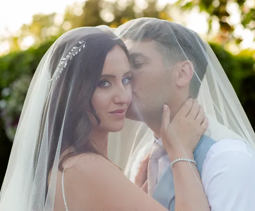 A bride and groom share a tender moment beneath a shared wedding veil outdoors in natural sunlight.