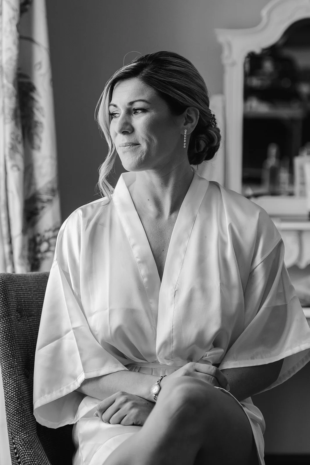 A black and white portrait of a bride getting ready, wearing a satin robe and delicate earrings while looking out a window.
