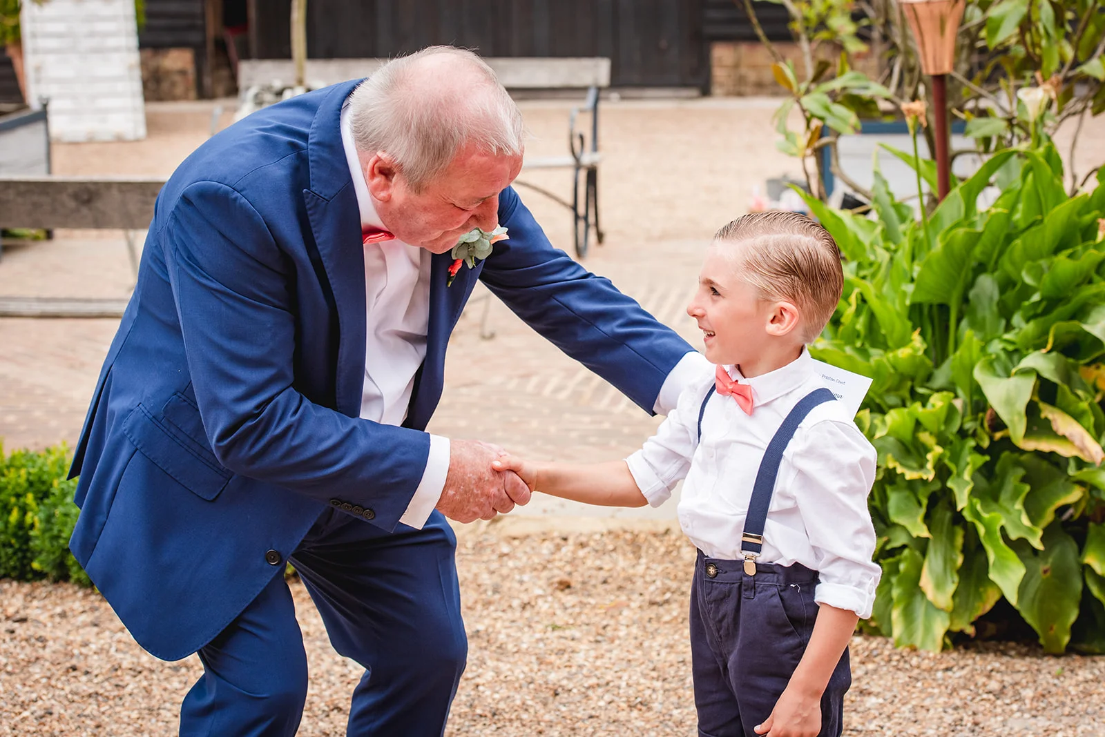 An older gentleman in a navy suit warmly shakes hands and smiles at a young page boy wearing matching trousers, braces, and a pink bow tie outdoors in a courtyard.