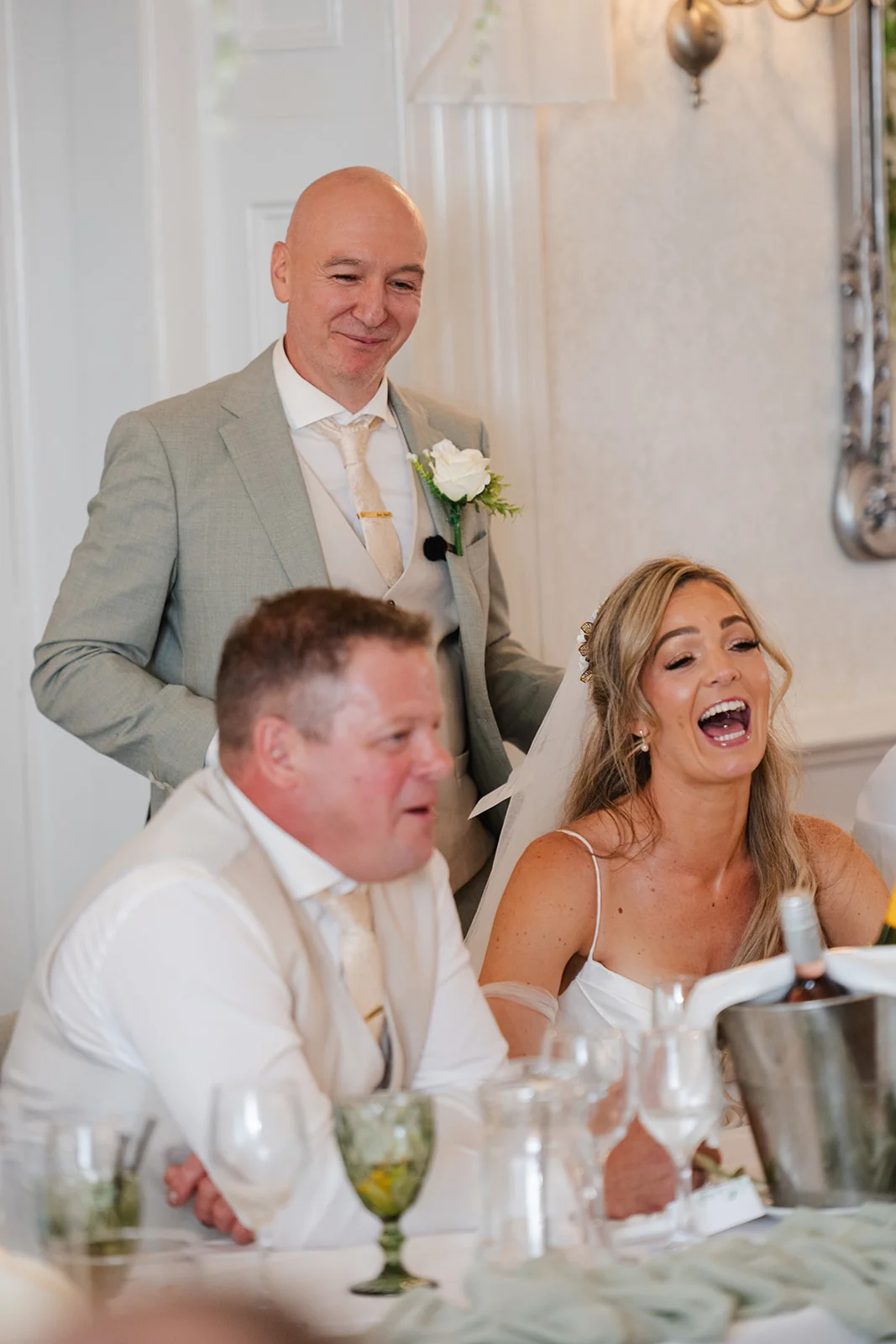 A bride throws her head back in a joyous belly laugh during the wedding speeches while seated at the top table next to the groom, with a guest in a grey suit looking on smiling.