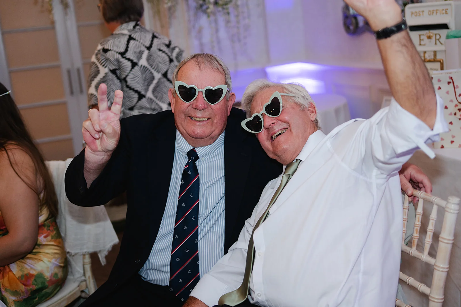 Two older male wedding guests wearing playful heart-shaped sunglasses smile and laugh while posing together at the reception.