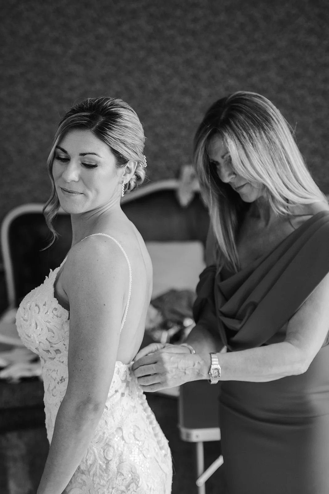 A mother helps her smiling bride daughter zip up her detailed lace wedding dress during morning preparations.