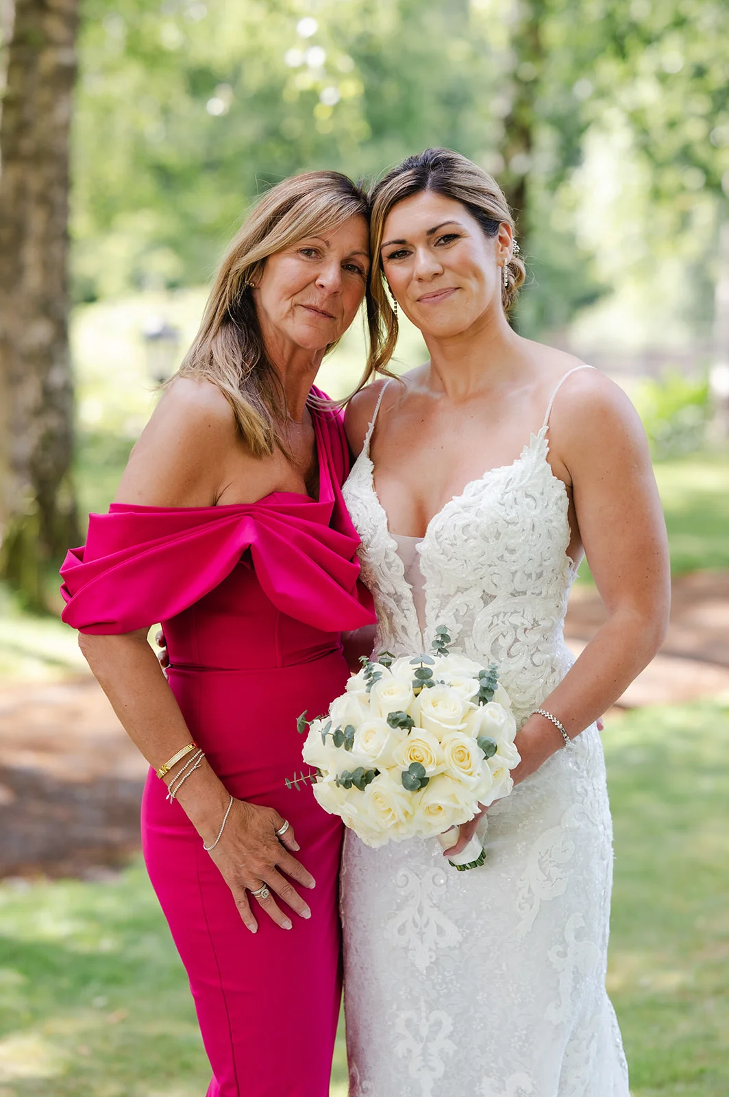 The bride in a lace gown with spaghetti straps and her mother in a bright pink dress pose together outdoors holding a bouquet of white roses.