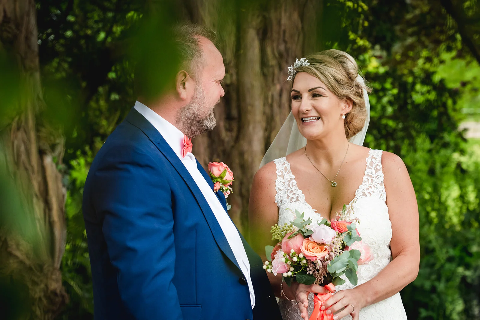 A smiling bride holding a colourful floral bouquet gazes lovingly at her groom in a blue suit amidst a lush green outdoor setting.