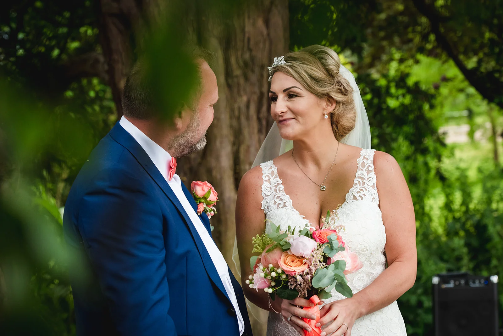 A smiling bride and groom gaze lovingly at each other outdoors, surrounded by lush greenery during their wedding ceremony.