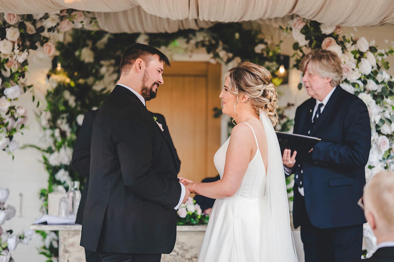 A bride and groom lovingly hold hands during an outdoor wedding ceremony beneath a magnificent white floral arch.
