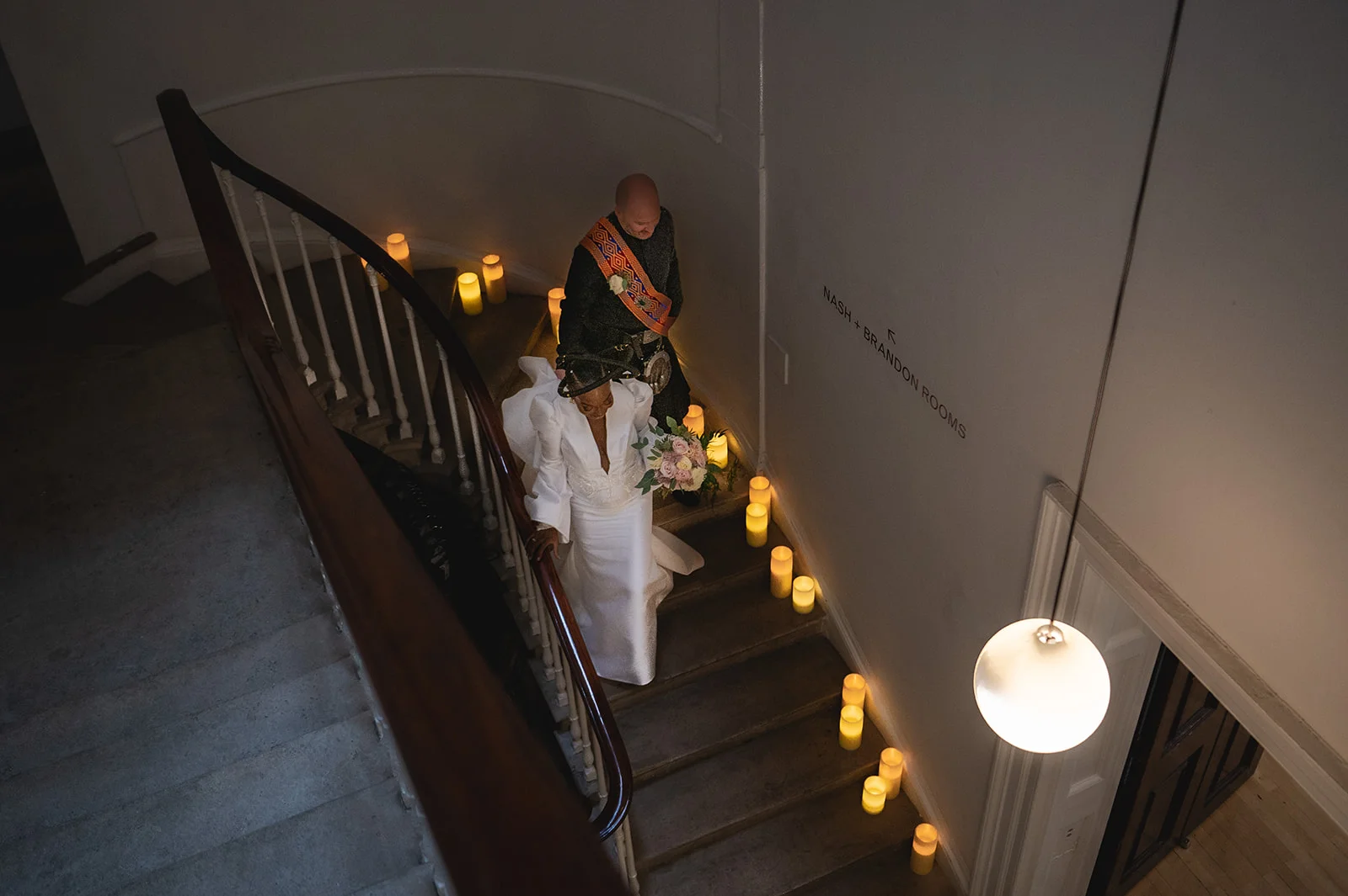 An interracial couple descends a grand candlelit spiral staircase, with the bride holding a delicate floral bouquet while wearing a modern white gown.