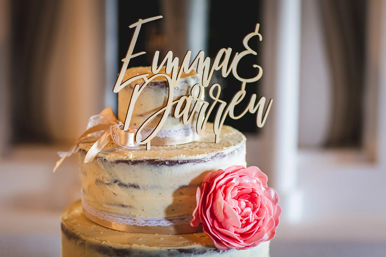 A close up view of a three-tier semi naked wedding cake decorated with a pink peony flower, lace ribbon, and a custom wooden name cake topper.