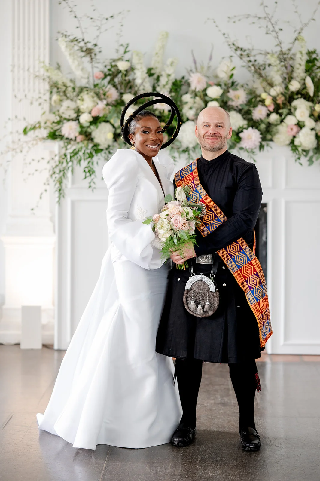 A newlywed couple poses in front of a white fireplace adorned with a lush floral installation, the bride wearing a striking white gown and sculptural halo headpiece while holding a bouquet, and the groom dressed in a black kilt and tartan sash.