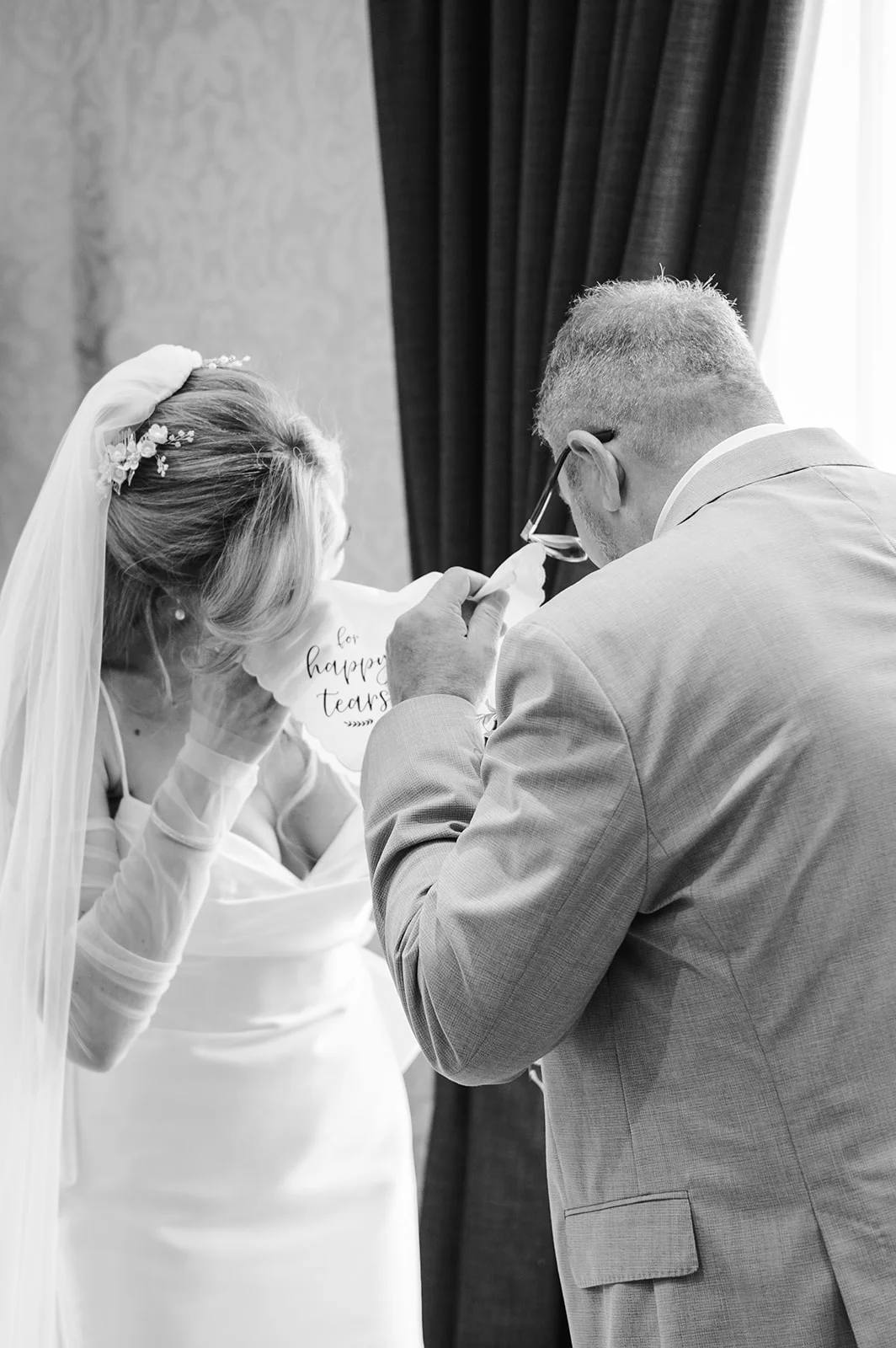 An emotional black and white shot of a tearful bride and her father sharing a tender moment while wiping away happy tears with an embroidered handkerchief.