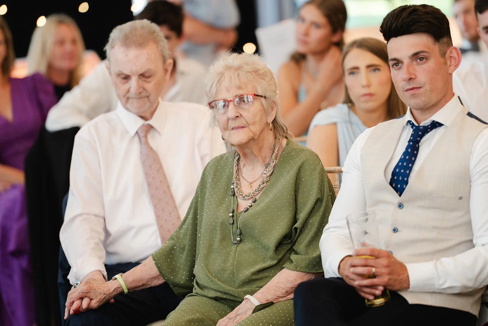 An older woman wearing glasses and an olive green top sits attentively amongst wedding guests, holding hands with a family member seated beside her.